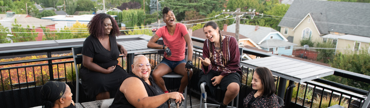 Overhead shot of six disabled people at a rooftop deck party. A person with a prosthetic leg smiles directly at the camera and gives a thumbs up while everyone else is engaged in conversation.