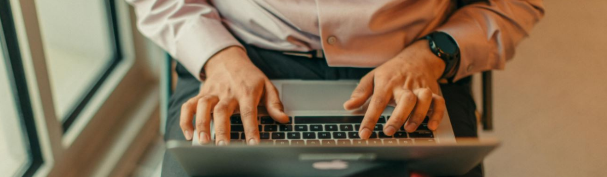 A close up shot of a person’s arms and hands as they type on their laptop. The person is sat down on a mobility scooter and is wearing dark smart trousers and light purple shirt.