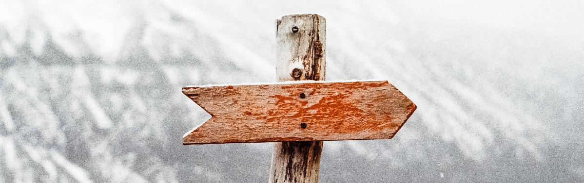 The top part of a simple, rustic, outdoor signpost made of a vertical wooden log, with a horizontal piece of wood in the shape of an arrow nailed into it.  The arrow points to the right and the sign is weathered and old.  It is set against an all-white background. 