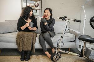 Two disabled women chat and sit on a couch, both holding coffee mugs. An electric lightweight mobility scooter rests on the side.